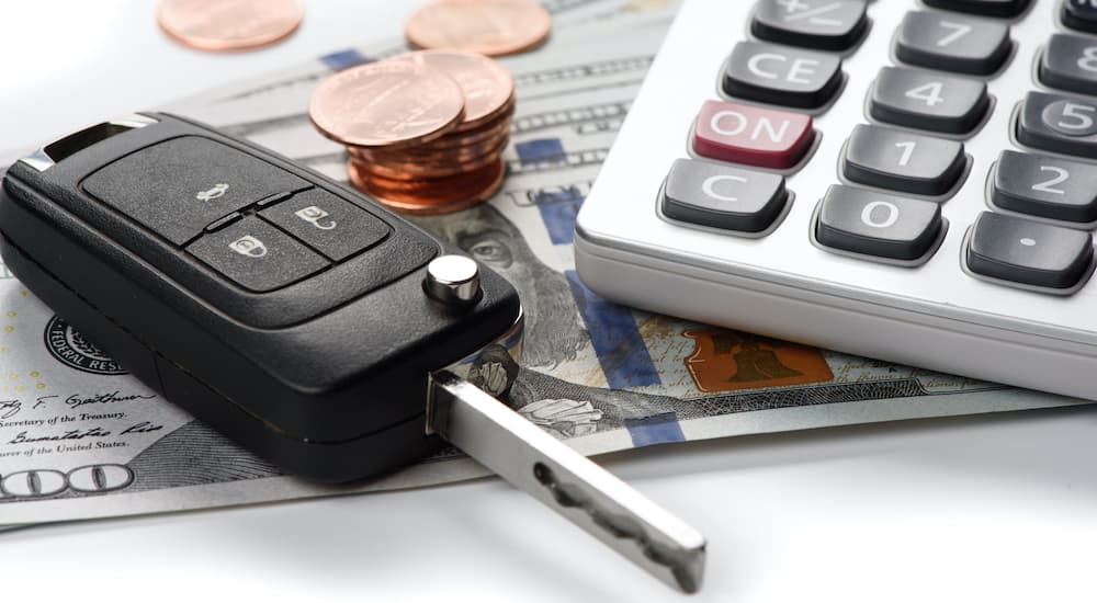 A car key is shown sitting on top of hundred dollar bills and a calculator.