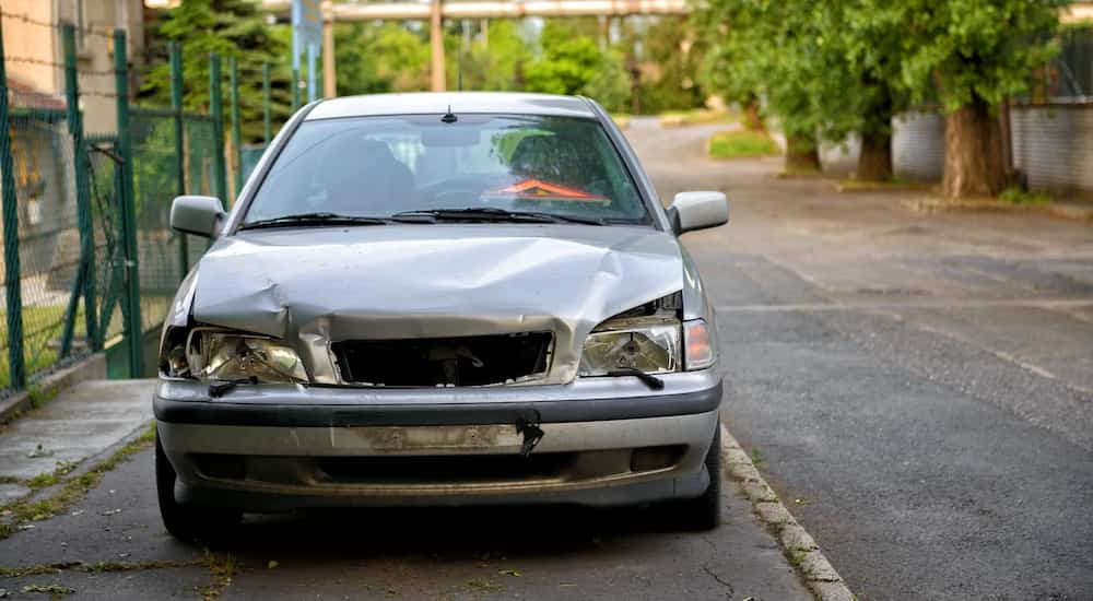 A silver beat up car is shown from the front parked on a city street after the owner searched 'sell my car'.