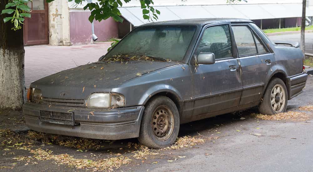 A dark grey car in poor condition is shown parked on a city street.