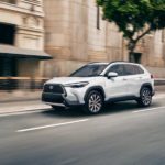 A white 2022 Toyota Corolla Cross is shown from the front at an angle while driving down a city street after the owner searched 'Toyota dealer near me'.