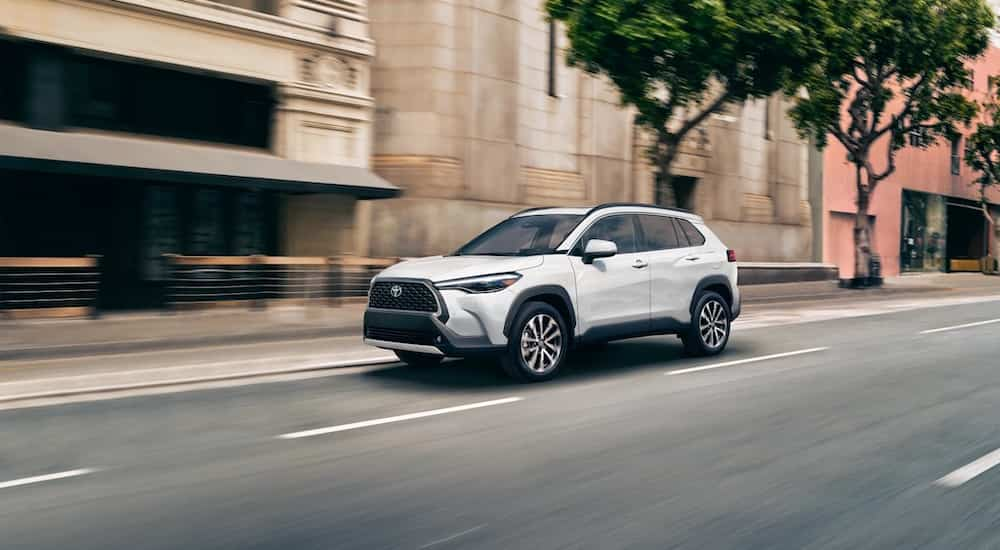 A white 2022 Toyota Corolla Cross is shown from the front at an angle while driving down a city street after the owner searched 'Toyota dealer near me'.