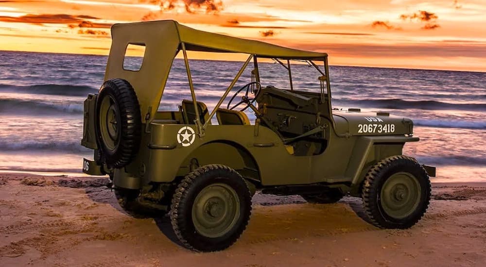 A green 1942 Jeep Wills is shown from the side parked on a beach at sunset.