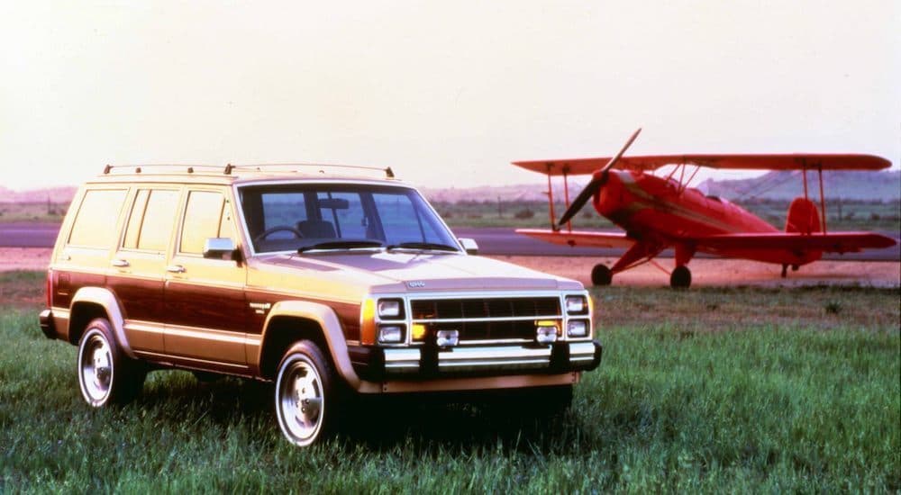 A brown 1984 Jeep Wagoneer is shown parked in a field next to a red plane.