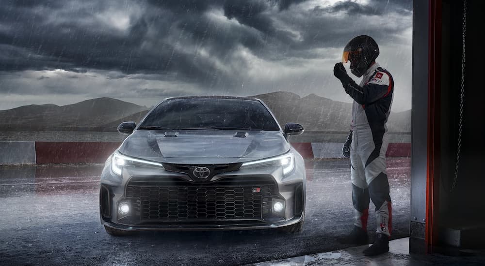 A grey 2023 Toyota GR Corolla is shown on a stormy day next to a race car driver.