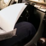 A person is shown sitting in the front seat of their car looking at paperwork.