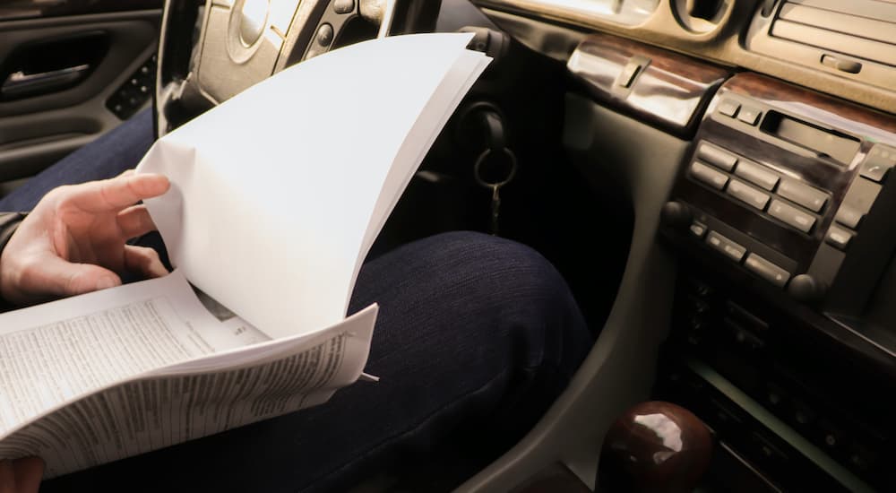 A person is shown sitting in the front seat of their car looking at paperwork.
