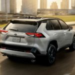 A silver 2020 Toyota RAV4 is shown from the rear driving past a city.