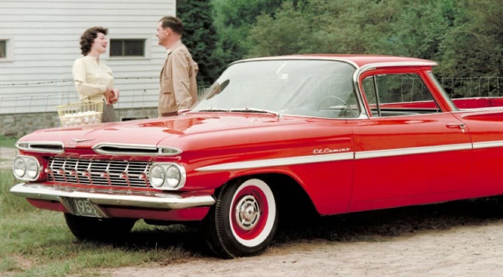 A couple is shown standing next to a red 1959 Chevy El Camino.