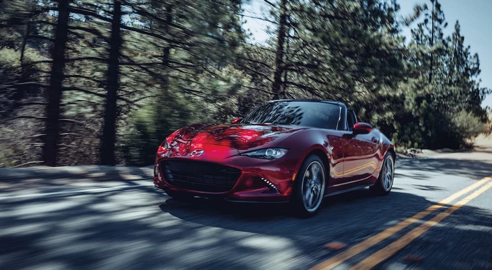A red 2022 Mazda MX-5 Miata is shown driving on a tree-lined road.