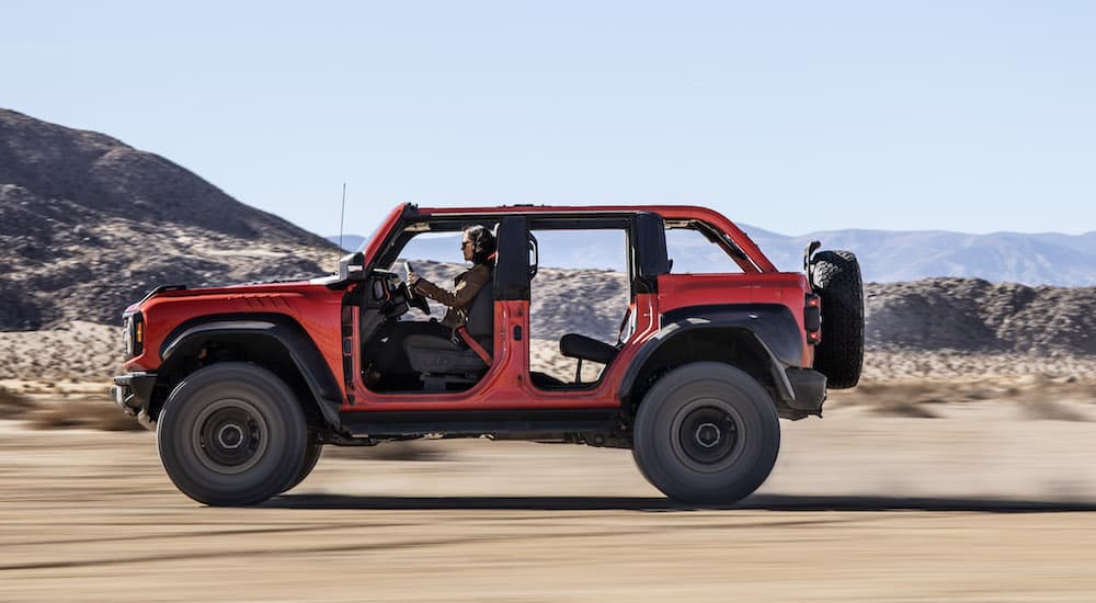 An orange 2022 Ford Bronco Raptor is shown from the side with no roof and doors driving through a desert.