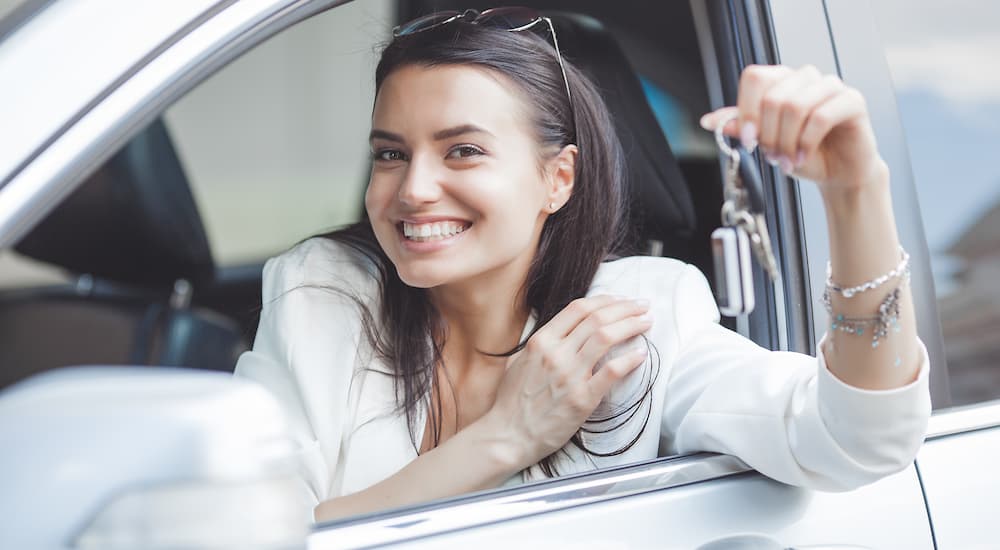 A woman is shown holding a car key out of a car window.