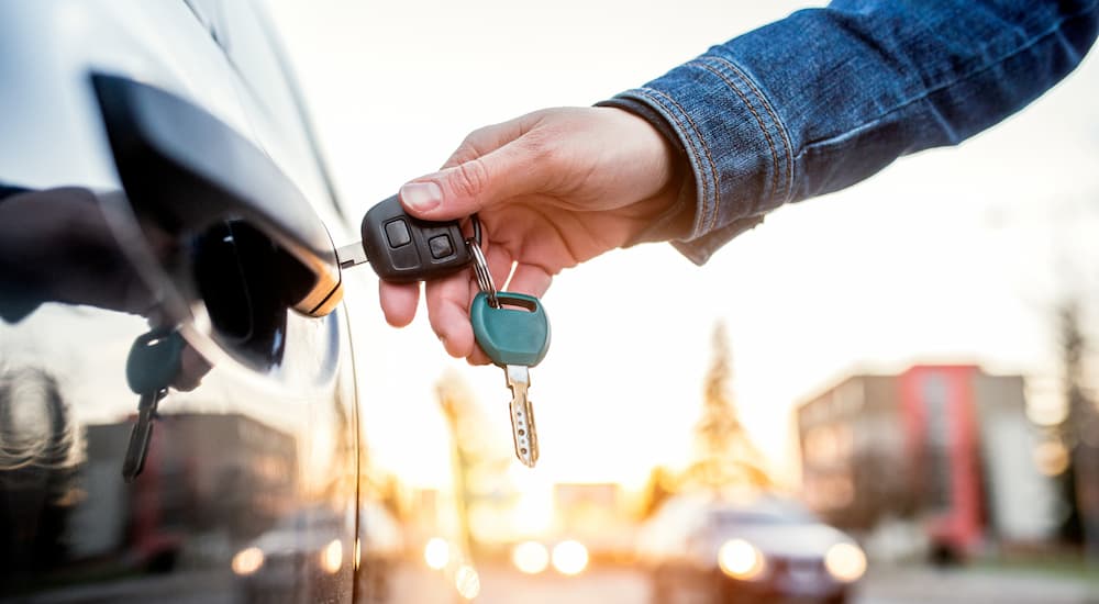 A person is shown using a car key to open a car door at a used car lot.