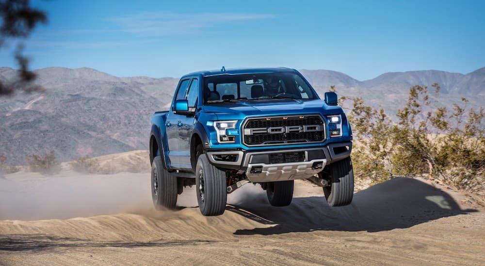 A blue 2019 Ford F-150 Raptor is shown from the front while jumping after leaving a dealer that advertised having used lifted trucks for sale..