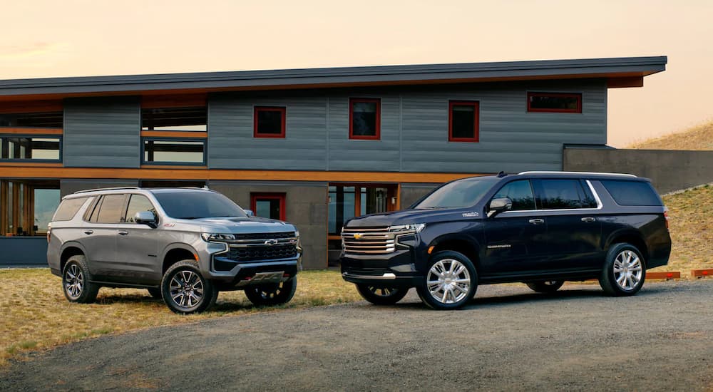 A silver 2023 Chevy Suburban Z71 and a dark blue High Country are shown facing each other in front of a house.