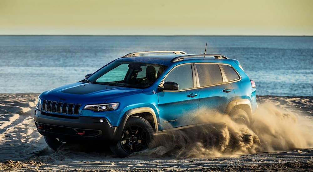 A blue 2022 Jeep Cherokee Trailhawk is shown from the side while kicking up sand on a beach.