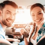 A couple is shown smiling in the front seat of a their vehicle.