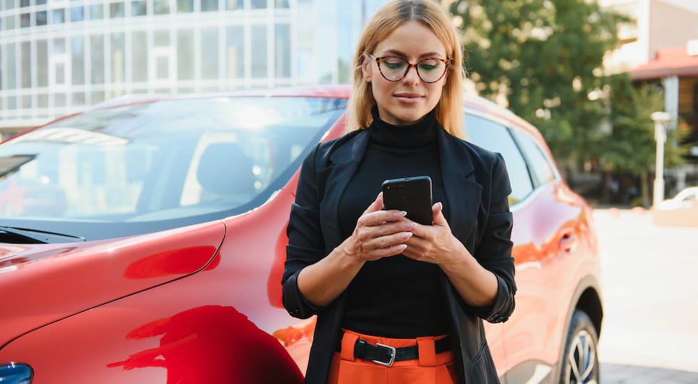 A woman is shown on her phone while standing next to a red vehicle.