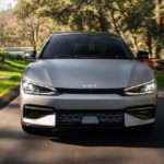 A silver 2023 Kia EV6 is shown from the front speeding down a tree-lined road.