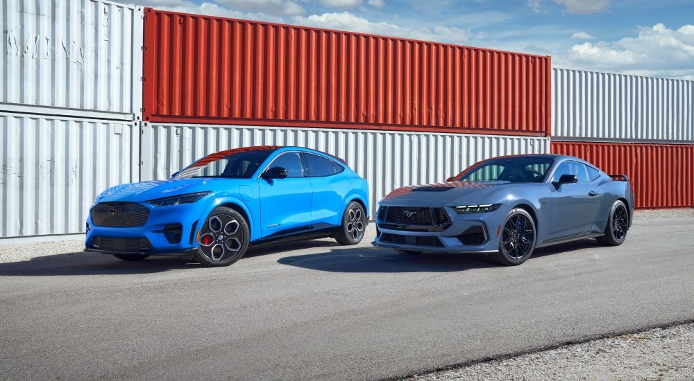 A blue 2024 Ford Mustang Mach-E and a dark blue 2024 Ford Mustang is shown parked near shipping containers.