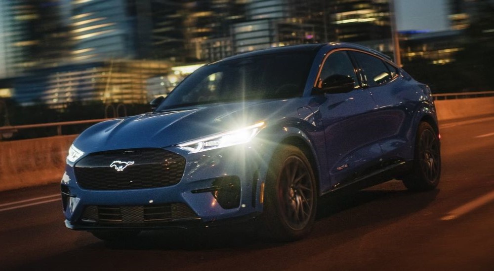 A blue 2024 Ford Mustang Mach-E for sale is shown driving in a city.