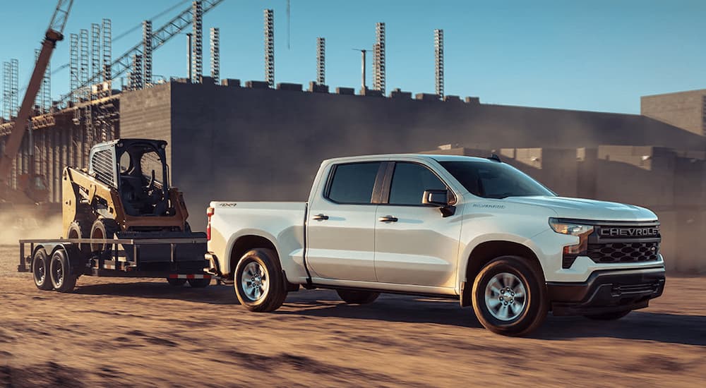 A white 2023 Chevy Silverado 1500 WT is shown towing heavy machinery through a construction site.
