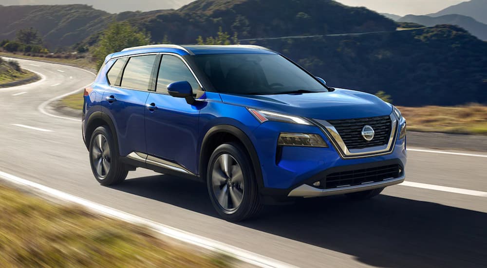 A blue 2021 Nissan Rogue for sale is shown driving on a winding road.