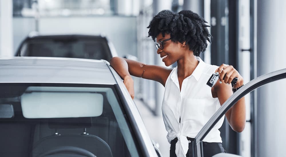 A woman is shown standing next to the open door of a white sedan.