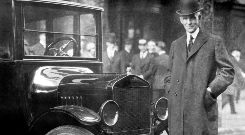 A black and white image of Henry Ford is shown standing next to a Model T.