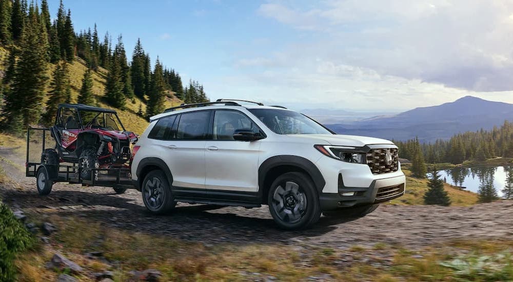 A white 2023 Honda Passport Elite is shown towing UTVs off-road.