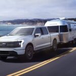 A white 2023 Ford F-150 Lightning is shown towing an Airstream trailer on a coastal road.