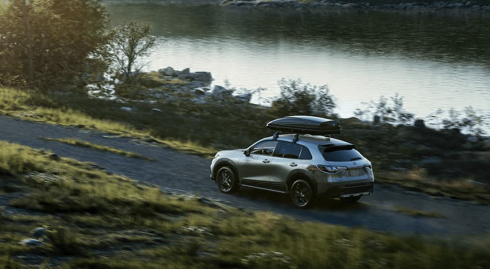 A silver 2023 Honda HR-V is shown from the rear driving on a dirt road next to a lake.