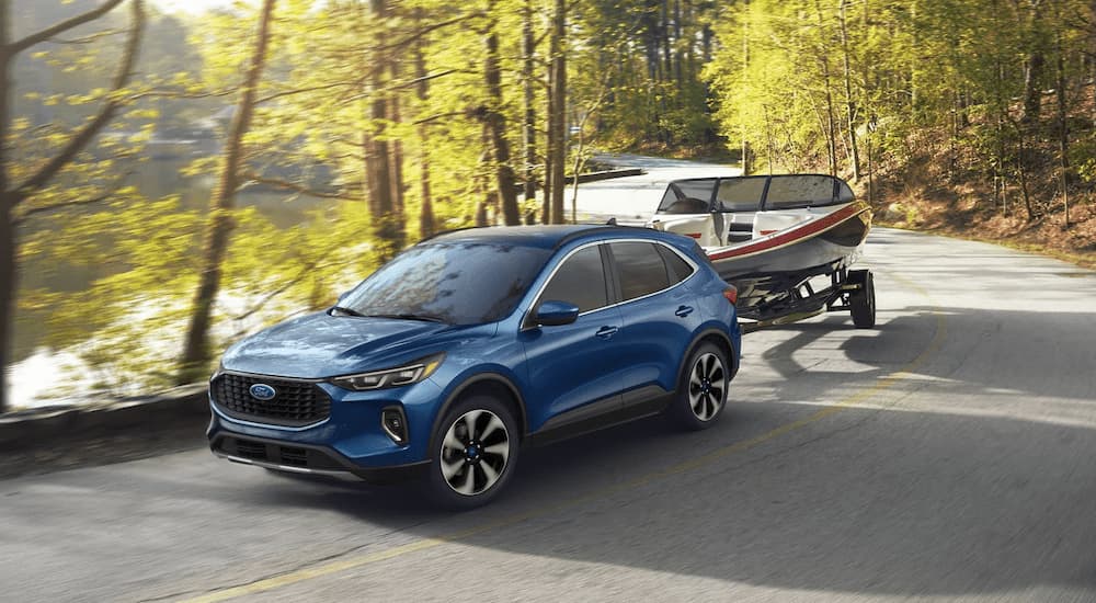 A blue 2023 Ford Escape is shown towing a small boat on a tree-lined road.
