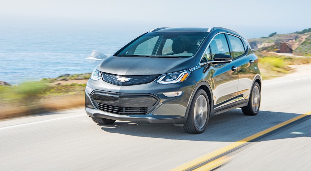 A grey 2019 Chevy Bolt EV is shown driving on an open road near an ocean.