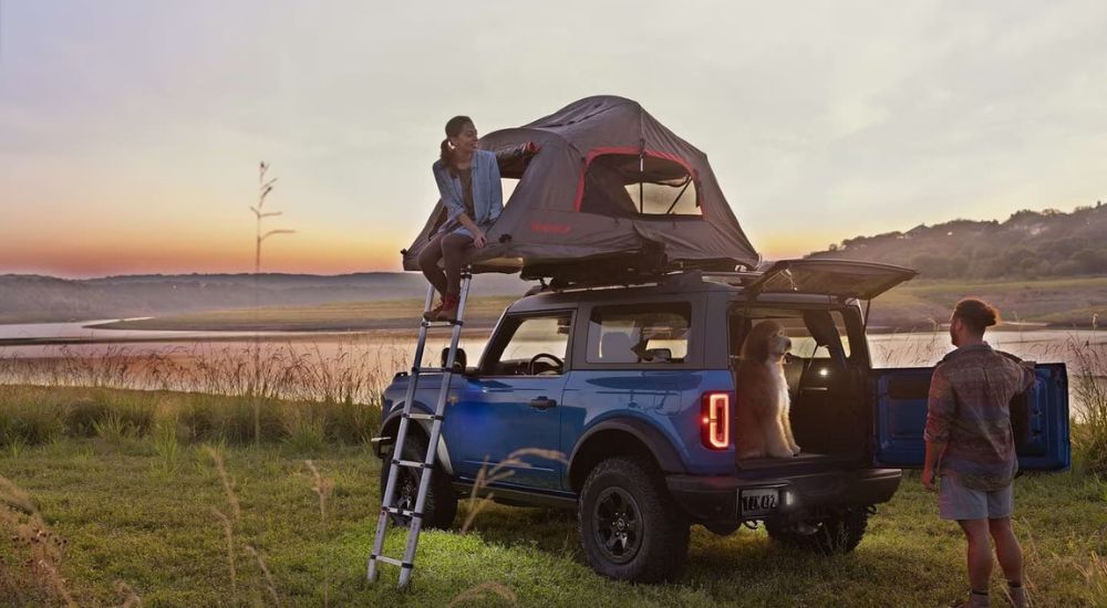 A blue 2023 Ford Bronco is shown with a woman sitting in a set up rooftop tent.