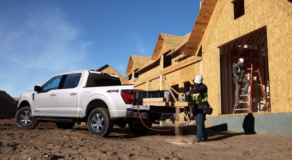 A white 2023 Ford F-150 is shown parked in front of a house being constructed while a worker uses the tailgate workspace to cut wood. 