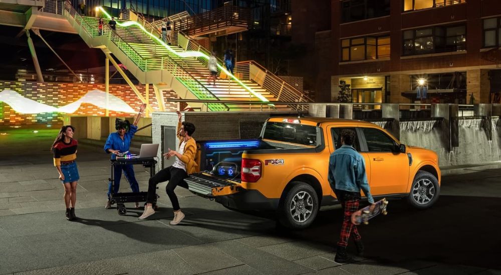 An orange 2023 Ford Maverick is shown parked in front of a lighted staircase while a group of friends have a tailgate party.
