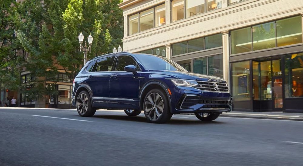 A blue 2023 Volkswagen Tiguan is shown driving on a street near city stores.