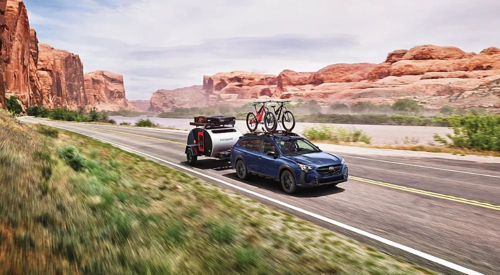 A blue 2023 Subaru Outback is shown driving down the road towing a trailer with bikes in a roof rack.