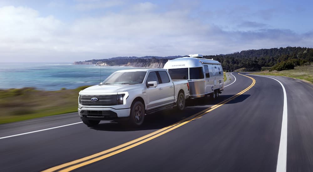 A white 2023 Ford F-150 Lightning is shown from the front at an angle while towing a camper.