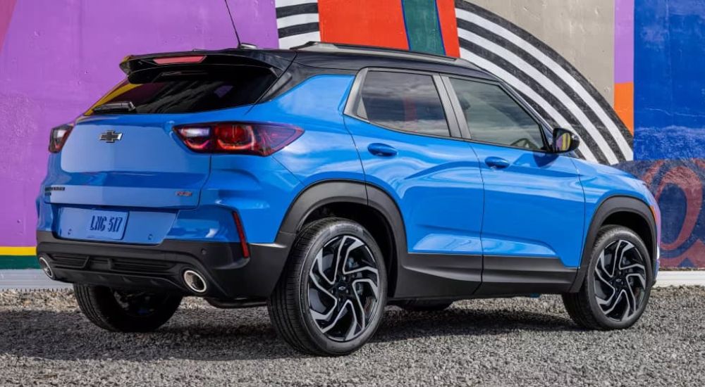 A blue 2024 Chevy Trailblazer is shown at an angle from the back parked in front of a wall with colorful graffiti. 