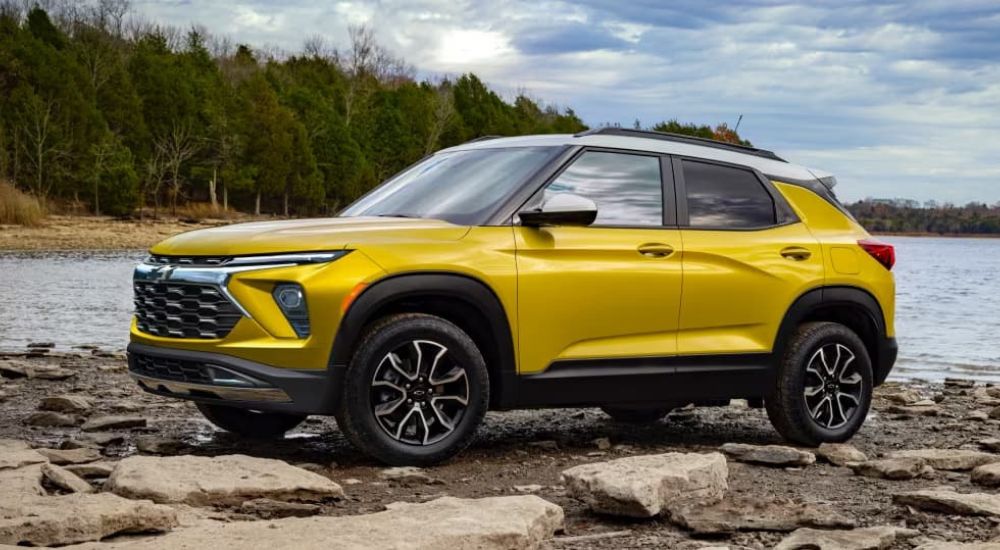 A yellow 2024 Chevy Trailblazer is shown on the rocky shore of a lake in the woods.