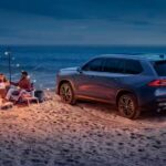 A family is shown gathered around a campfire on a beach beside a blue 2024 Toyota Grand Highlander.