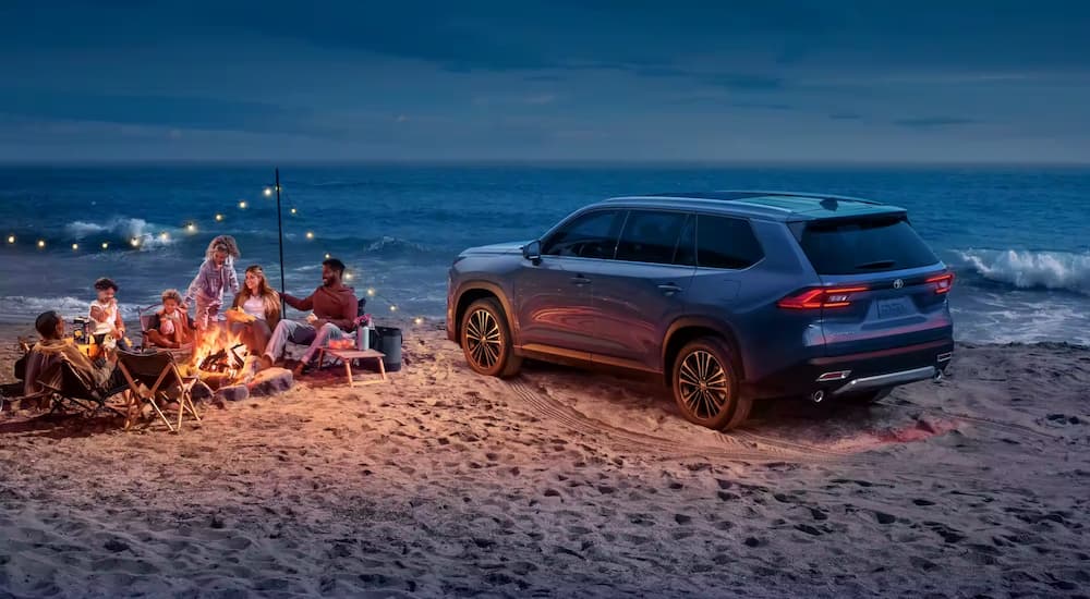 A family is shown gathered around a campfire on a beach beside a blue 2024 Toyota Grand Highlander.