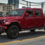 A red 2023 Jeep Gladiator for sale is shown driving on a city bridge.