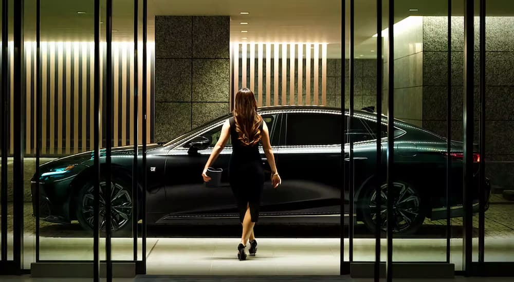 A woman in black dress walks towards a black 2023 Lexus LS 500 F Sport parked outside a modern building.