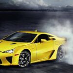 A yellow 2012 Lexus LFA is shown emerging from a cloud of smoke in an empty parking lot.