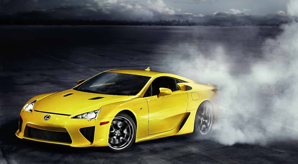 A yellow 2012 Lexus LFA is shown emerging from a cloud of smoke in an empty parking lot.