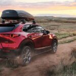 A red 2023 Mazda CX-30 with a roof box is shown driving down a dirt road across the prairie.