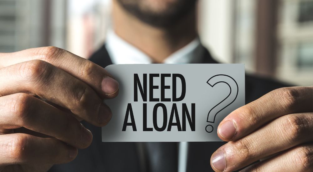 A close up shot of the hands of a business man holding a paper that reads "Need a Loan?" is shown.