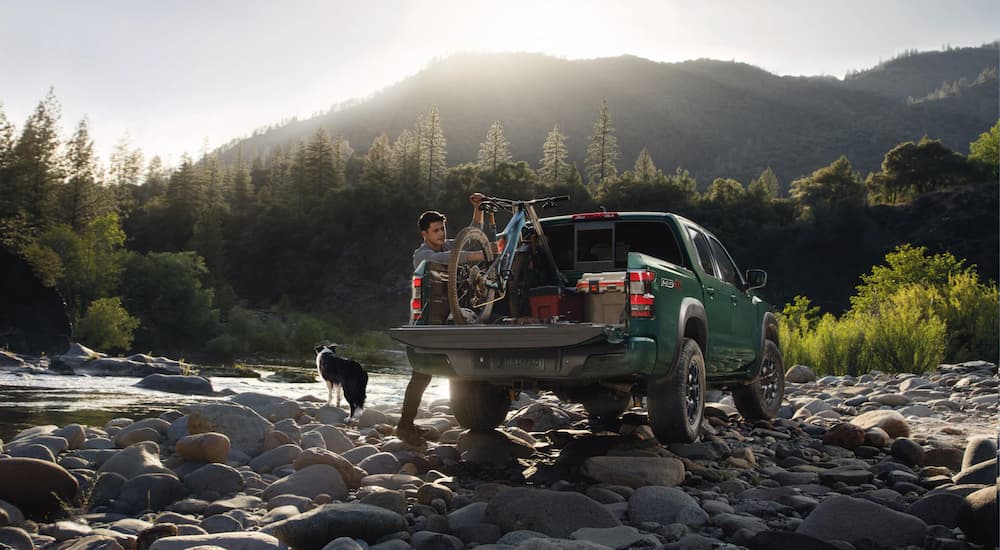 A man is shown unloading a bicycle from a green 2023 Nissan Frontier PRO-4X parked beside a forest creek as a black and white dog stands nearby.